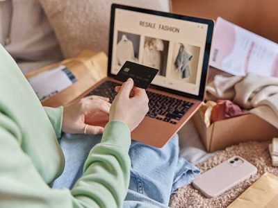 Pessoa segurando cartão de crédito preto em frente a um notebook aberto em site de comércio eletrônico de roupas, cercada por encomendas. Local da imagem: Tráfego Pago Google Ads - Menu DT